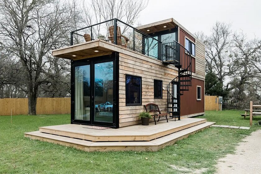 two floors brown and wooden Shipping Container Home with Glassdoor and small Waking area and blac rotating Stair going to second floor sitting area.