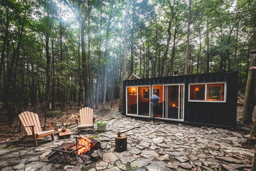 Black container home Cabin yellow lights inside one hall connected glass door and one bedroom on other side, camp fire outside 